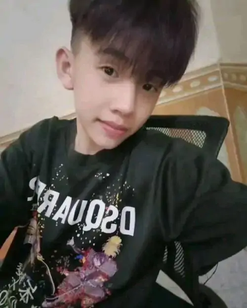 Trai đẹp 2k11 mặc áo đen in chữ đứng trong phòng, tóc nâu phủ trán và biểu cảm hơi nghiêng tạo nét dễ gần