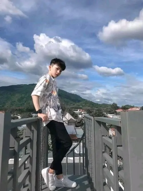 Trên lan can cầu giữa cảnh núi biển, bạn nam áo sơ mi xám tạo dáng ngồi, tóc rối nhẹ và ánh sáng ngoài trời rất trong