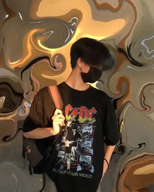 Ánh đèn vàng phản chiếu trên tường loang màu, cậu bạn Gen Z mặc áo đen đứng che mặt, vòng cổ bạc tạo điểm nhấn