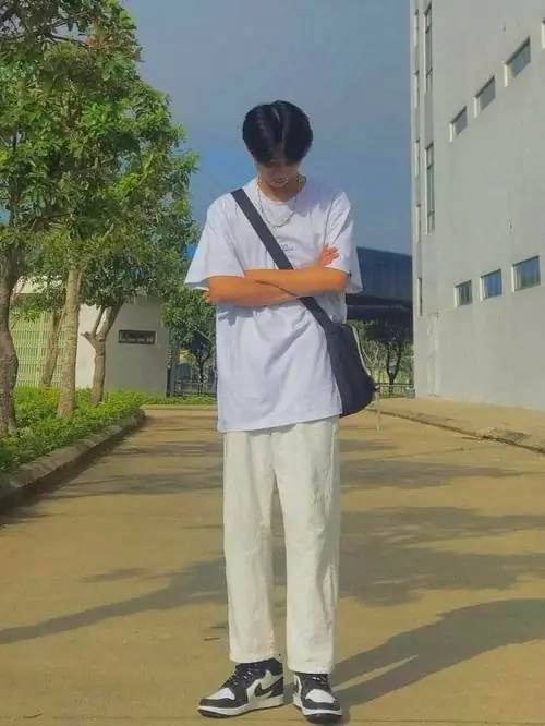 Bạn nam mặc áo phông trắng và quần sáng màu đứng khoanh tay bên tòa nhà, kiểu tóc gọn làm dáng vẻ thêm nhẹ nhàng