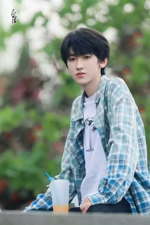 Giữa nền hoa lá mờ, chàng trai áo kẻ xanh ngồi nhìn thẳng, mái tóc đen mềm và gương mặt sáng tạo cảm giác thư sinh