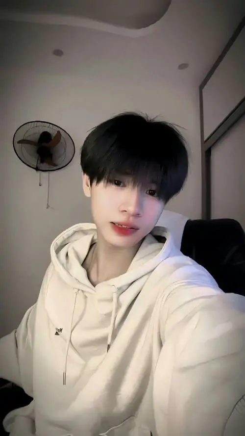 Hoodie kem kéo khóa làm cậu bạn ngồi trên giường trông ấm áp, mái tóc đen dày và biểu cảm yên lặng rất ngoan