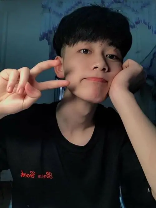 Hai ngón tay chạm má khiến bạn nam K13 áo đen trông đáng yêu, mái tóc gọn và nền xanh tạo cảm giác mát