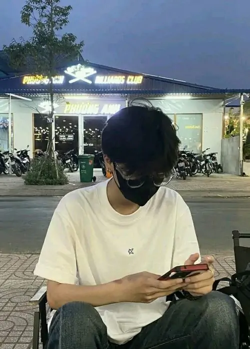 Sân trường buổi tối làm bạn nam áo trắng ngồi xem điện thoại nổi bật hơn, tóc đen che trán và dáng rất thư thái