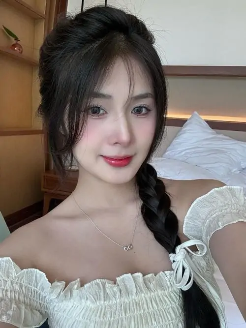 Áo trắng trễ vai làm cô gái tóc đen mỉm cười thêm dịu dàng, không gian phòng ngủ sáng nhẹ tạo cảm giác thân quen