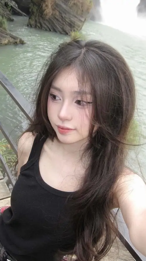Cô gái tóc đen mặc áo đen đứng trước rèm sáng, gương mặt nhỏ hơi nghiêng và ánh nhìn nhẹ tạo nét đời thường