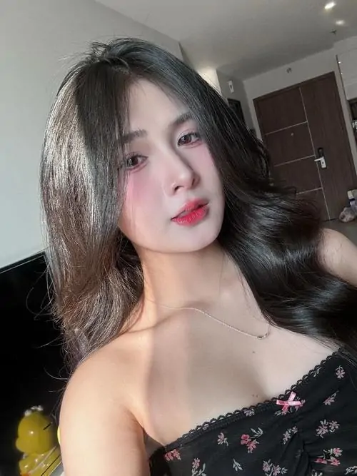 Cô gái tóc xoăn mặc váy trễ vai đen ngồi trong phòng, ánh sáng mềm giúp làn da và đường nét gương mặt hiện rõ
