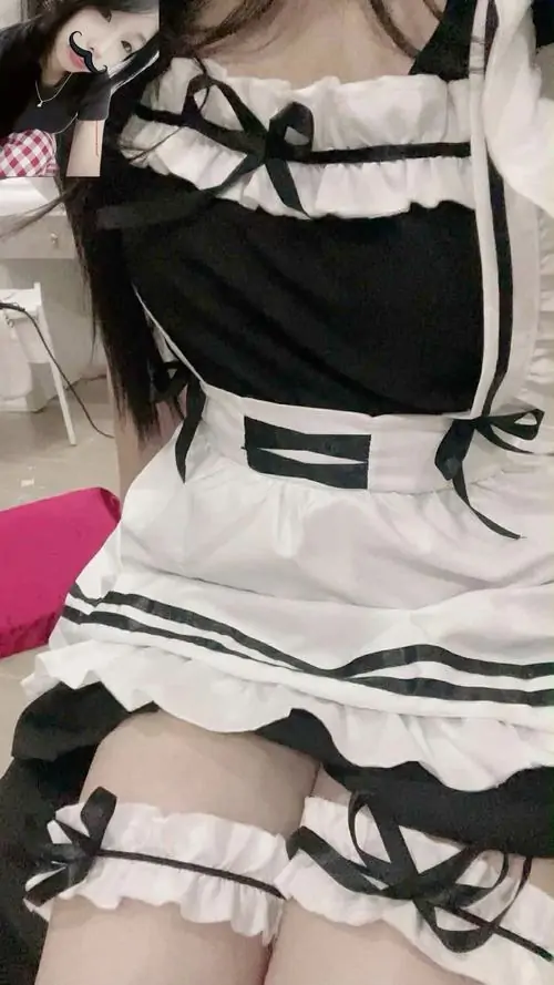 Cô gái ngồi cạnh bàn gỗ, đôi bàn tay nhỏ nhắn đặt lên gối, trang phục hầu gái sạch sẽ tạo cảm giác trong sáng
