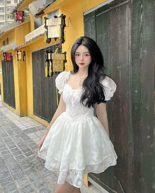 Trước dãy nhà màu vàng, cô gái váy trắng ren đứng thẳng, tóc đen uốn sóng và túi nhỏ đeo bên vai