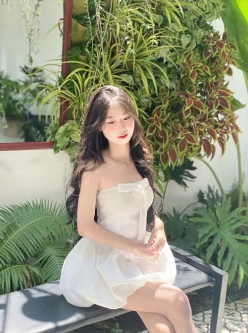 Góc vườn nhiều cây xanh làm cô nàng váy trắng hai dây thêm mềm, dáng ngồi khép chân và biểu cảm hơi trầm
