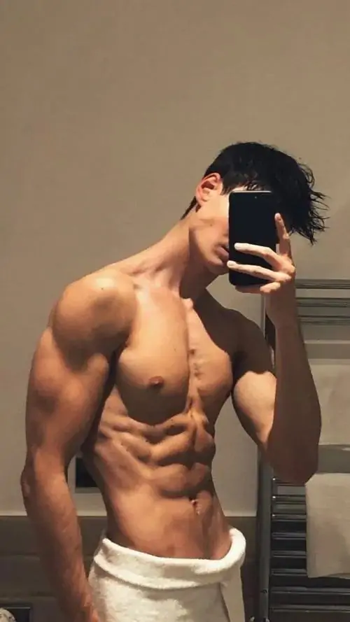 Phòng tập sáng đèn phản chiếu trên gương lớn, nam mẫu cầm chai nước, tóc ướt nhẹ và cơ bụng hiện sau áo mở khóa