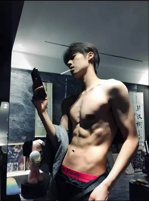 Trong phòng gym ánh sáng mạnh, chàng trai tóc đen quay nghiêng, mặc quần tối, cơ bụng rõ nét và ánh mắt nhìn sang bên