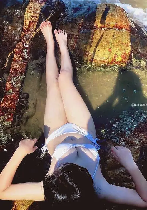 Bên bãi biển mùa hè, cô gái mặc áo khoác mỏng ngoài bikini, tóc bay nhẹ và làn da khỏe nổi bật trong nắng