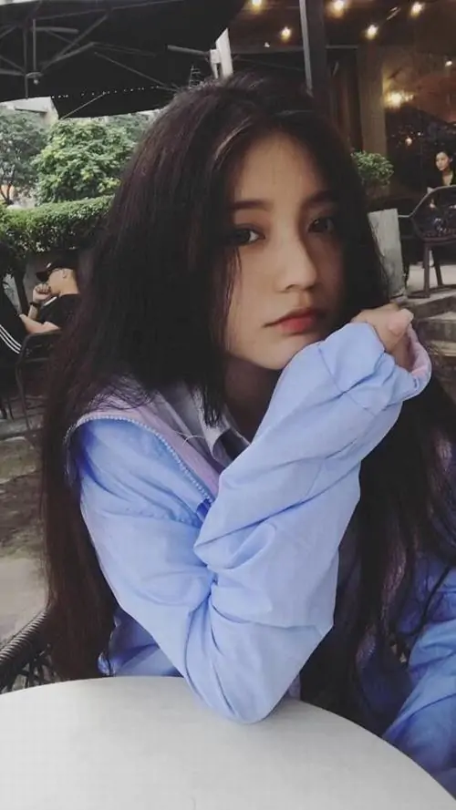 Cô gái mặc áo blazer kem ngồi trong quán sáng, tóc đen dài uốn nhẹ và gương mặt thanh tú tạo vẻ sang nhã
