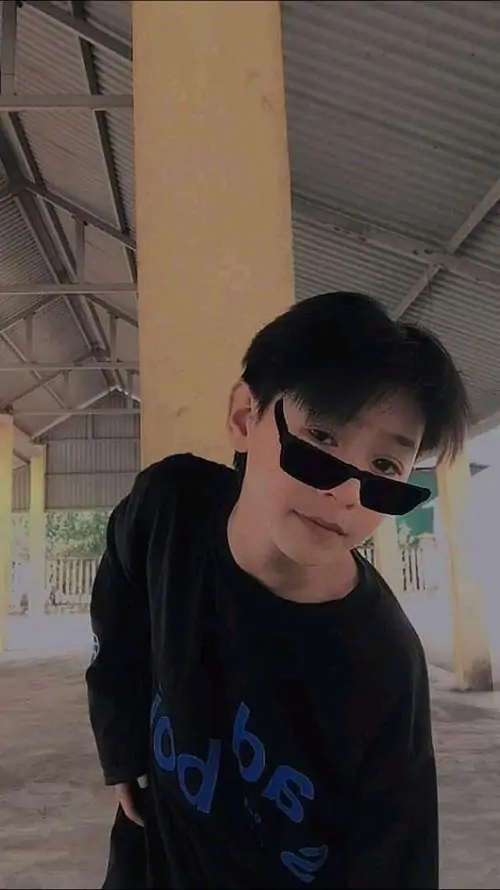 Bạn nam mặc đồng phục trắng đứng dưới hàng cây, tay cầm sách trước ngực, nụ cười hiền làm khung ảnh thêm sáng sủa