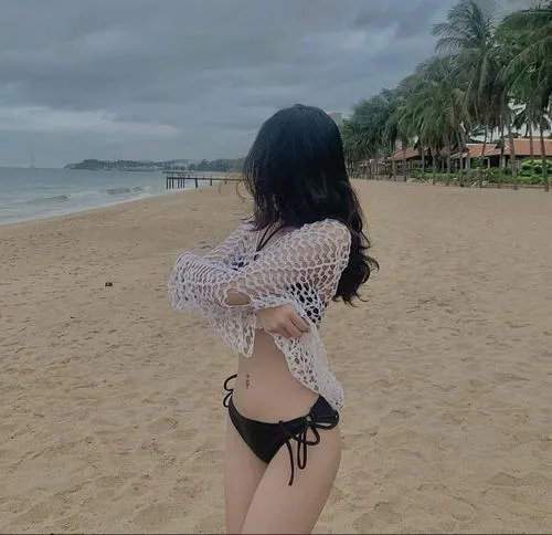 Ảnh người đẹp mặc bikini đỏ ngồi trên mỏm đá, mái tóc dài bay nhẹ và làn da sáng nổi bật giữa nền biển xanh