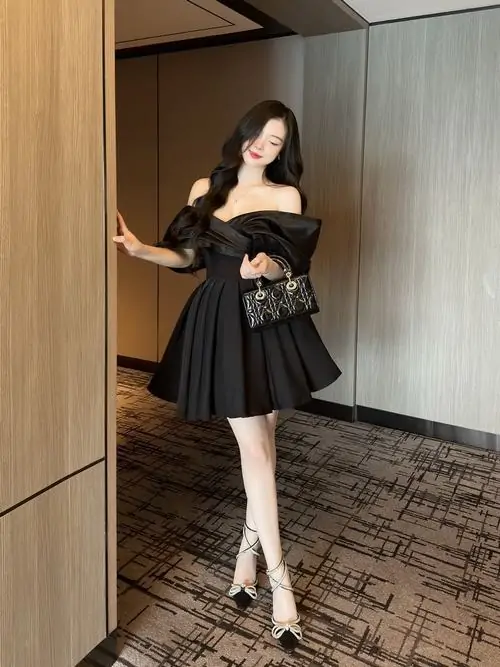 Cô gái diện váy đen xòe ngắn đứng cạnh cửa gỗ, đôi chân trắng nổi bật dưới ánh sáng trong nhà dịu mắt