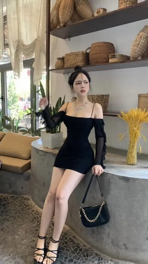 Ở góc phòng khách, cô nàng mặc váy đen ôm dáng ngồi trên ghế, tay cầm túi nhỏ và tạo dáng tự nhiên