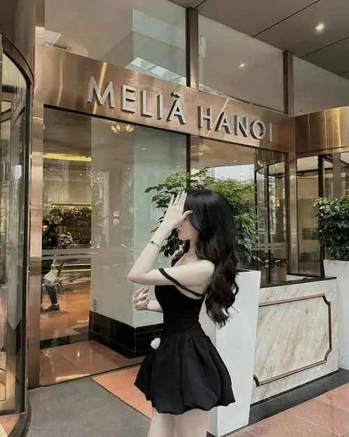 Gái xinh mặc váy ngắn đen đứng trước khách sạn, tay vuốt tóc nhẹ, phom váy xòe tạo cảm giác trẻ trung sang mắt