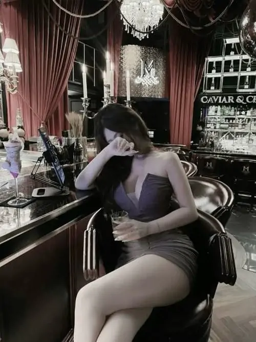 Bên khung cửa quán cà phê, cô gái mặc váy đen hai dây ngồi nghiêng, tay vuốt tóc và thần thái trầm nhẹ