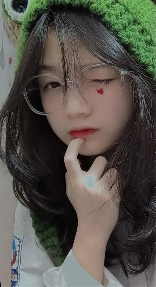 Nữ sinh đeo kính cận cài chiếc mũ len xanh tinh nghịch đang đưa ngón tay lên cằm tạo biểu cảm đáng yêu trước ống kính máy ảnh
