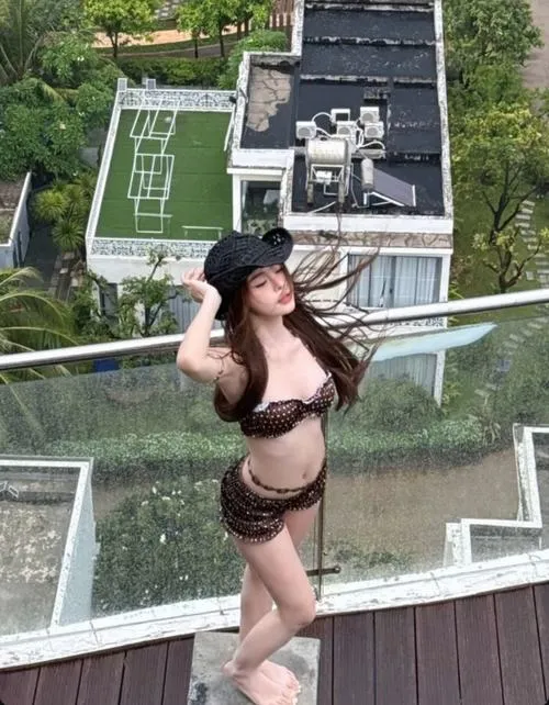 Người mẫu ngồi trong làn nước cạn với bikini vàng, mái tóc tím sẫm buông dài và khung biển mờ phía sau rất êm