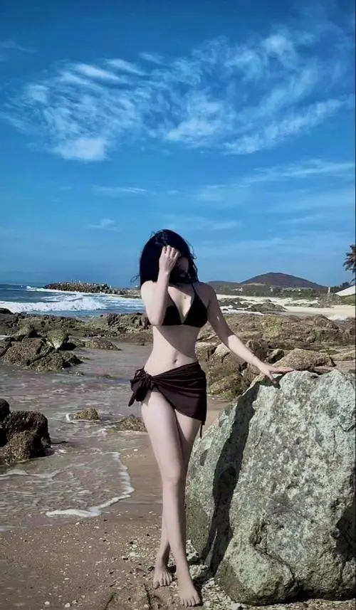 Giữa bầu trời xanh và bãi đá ven biển, cô gái mặc đồ bơi đen nghiêng người nhẹ, dáng mảnh mai tạo cảm giác năng động mùa hè
