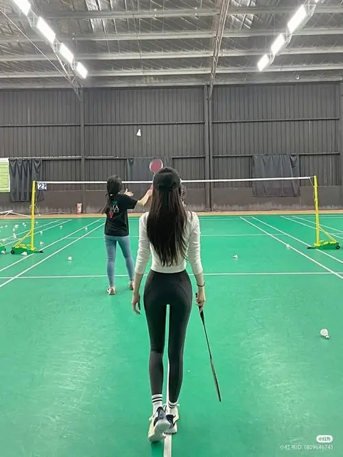 Cô gái quay lưng trên sân cầu lông xanh, tóc dài thả tự nhiên, áo thể thao trắng ôm gọn và quần legging đen nhìn khỏe khoắn