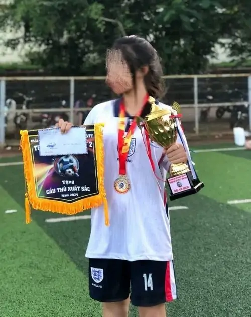 Cô gái đeo huy chương trên sân bóng, áo đấu trắng số mười bốn phủ ngoài, mái tóc ngang vai và phụ kiện cổ vũ nổi bật