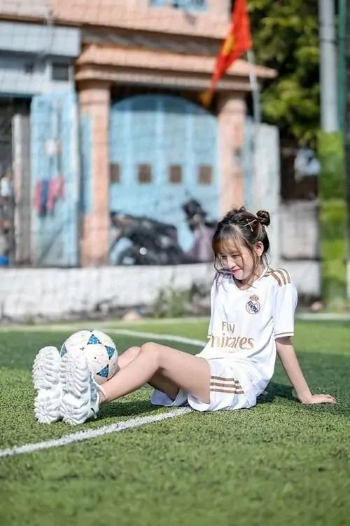 Bên quả bóng trắng đen, ảnh thiếu nữ cổ vũ bóng đá ngồi trên sân cỏ, áo đấu trắng và tóc búi hai bên nhìn rất tươi