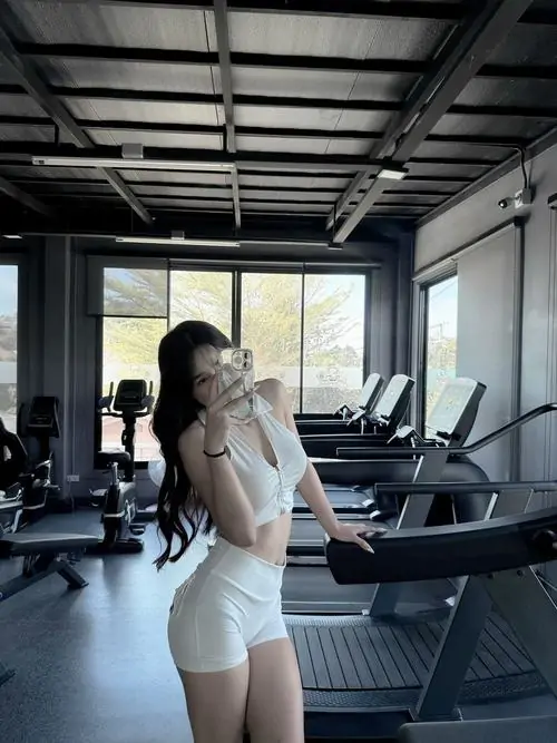 Trong phòng gym ánh sáng dịu, cô gái mặc set thể thao trắng đứng nghiêng, tóc dài thả lưng và vóc dáng thon gọn