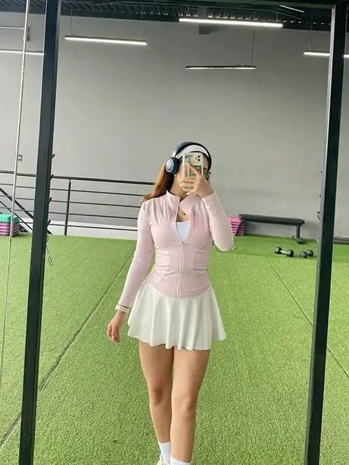 Cô gái mặc set tennis hồng nhạt đứng trên sân cỏ nhân tạo, tóc dài buông nhẹ, dáng tạo hình tự nhiên và tông ảnh sáng