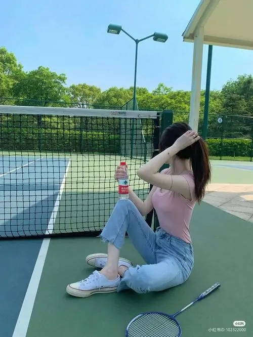 Dưới mái che sân tennis, cô gái ngồi nghỉ cạnh vợt và bóng, áo hồng phối quần jean tạo vibe thể thao đời thường