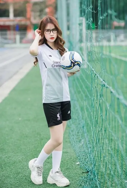 Cô gái cầm bóng trên sân cỏ, áo đấu trắng phối quần short đen, nụ cười nhẹ và mái tóc nâu tạo cảm giác năng động