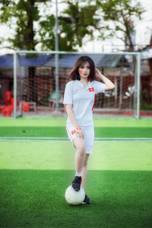 Giữa nền sân bóng xanh mướt, ảnh hot girl sân cỏ mặc áo thi đấu trắng, cầm quả bóng trước chân với dáng đứng tự tin
