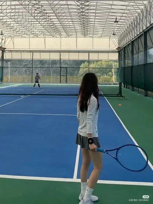 Cô gái quay lưng trên sân tennis xanh, áo thun trắng và quần short thể thao tạo dáng gọn gàng, tóc dài rơi tự nhiên