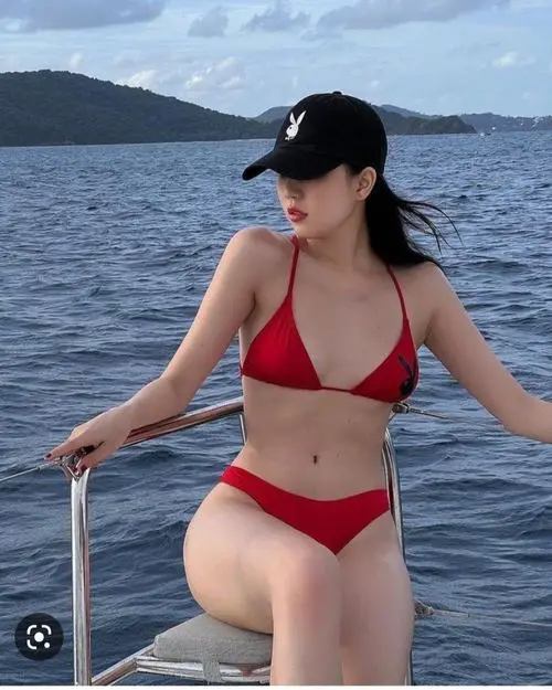 Bikini đỏ hai mảnh nổi bật trên nền biển xanh, người mẫu đội mũ đen đứng vịn lan can với dáng nghiêng gợi cảm nhẹ