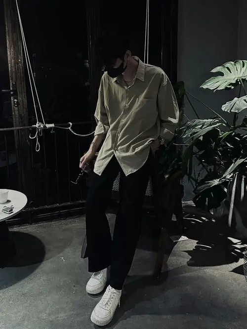 Bước đi thong thả của trai đẹp che mặt mặc chiếc áo khoác kaki sáng màu giữa khuôn viên có nhiều cây xanh xung quanh lối đi.