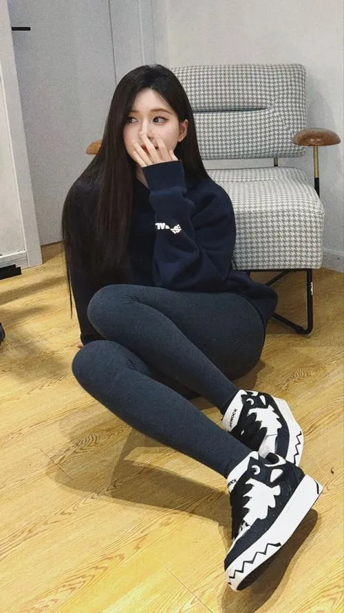 Ngồi trên sàn gỗ, cô gái mặc hoodie đen và legging tối màu che nhẹ khuôn mặt, giày thể thao cổ cao tạo điểm nhấn cá tính