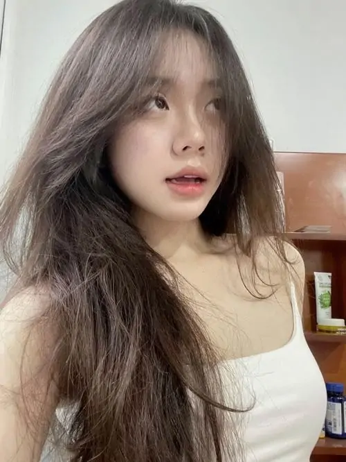 Gái xinh cười với mái tóc nâu hơi rối và áo kem đơn giản, góc chụp trong nhà làm ánh mắt trông thật tự nhiên