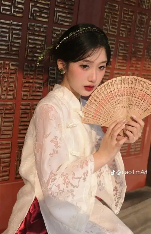Cận cảnh nàng áo dài trắng cầm quạt giấy trước ngực, gương mặt nghiêng nhẹ và ánh mắt mềm giữa nền gỗ tối