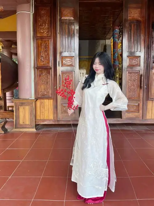 Cô gái áo dài trắng đứng giữa hiên gỗ đỏ, tay cầm hoa nhỏ và nhìn nghiêng trong không gian truyền thống ấm màu