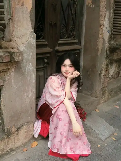 Ở góc cột đá cổ, cô gái áo dài hồng ngồi xổm cầm hoa, ánh mắt nhìn gần ống kính rất hiền