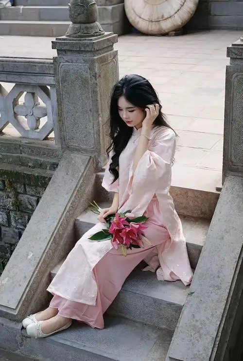 Trên bậc đá cổ, nàng áo dài hồng ngồi nghiêng người cầm hoa, ánh mắt nhìn xuống tạo dáng nhẹ trong khung hình dọc