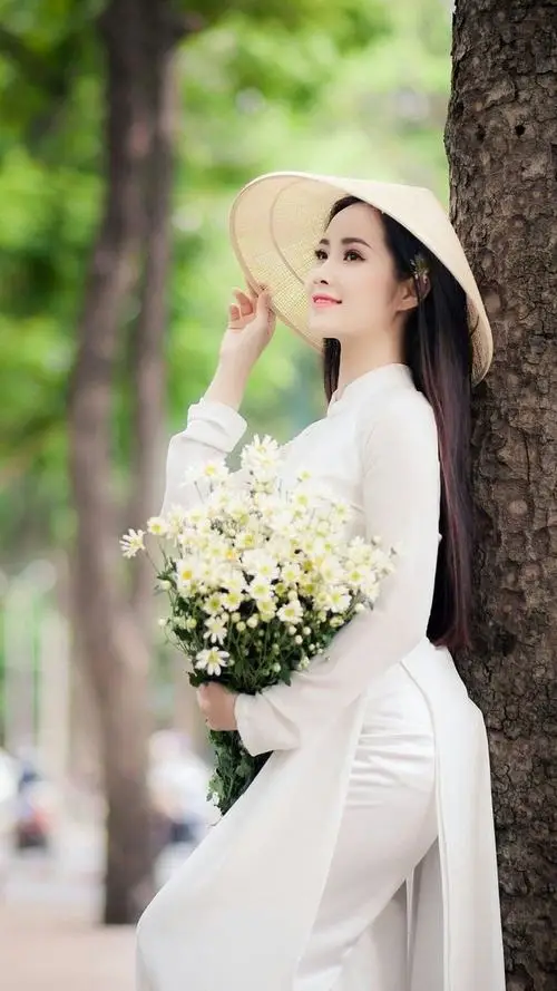 Giữa lối cây xanh, nàng áo dài trắng đội nón lá cầm bó hoa trắng, dáng đứng nghiêng tạo cảm giác thanh tao