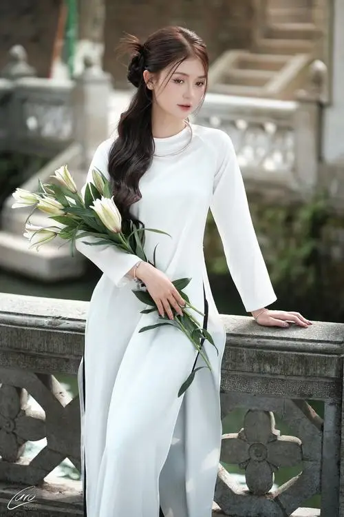 Bên lan can đá, cô gái áo dài trắng cầm cành sen, thân người xoay nghiêng để tà áo rơi dài mềm mại
