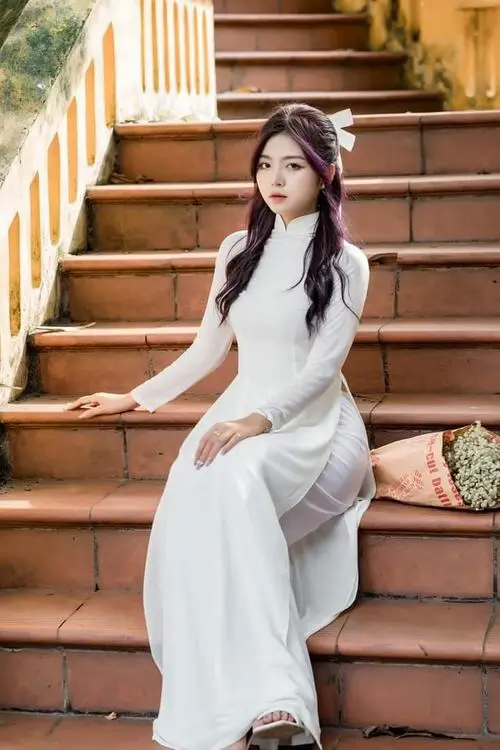 Trên cầu thang gỗ, cô gái áo dài trắng ngồi nghiêng người, một tay đặt lên bậc và nhìn thẳng đầy dịu dàng