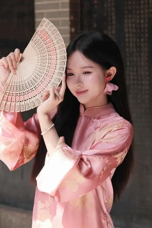 Cô gái áo dài hồng cầm quạt giấy cạnh má, gương mặt sáng và nụ cười nhẹ nổi bật trên nền gỗ tối