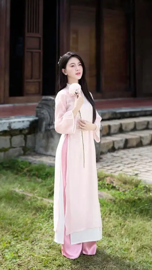 Nàng áo dài trắng đứng trước bậc thềm cũ, tay cầm hoa nhỏ bên hông theo tạo dáng đồ truyền thống thanh lịch