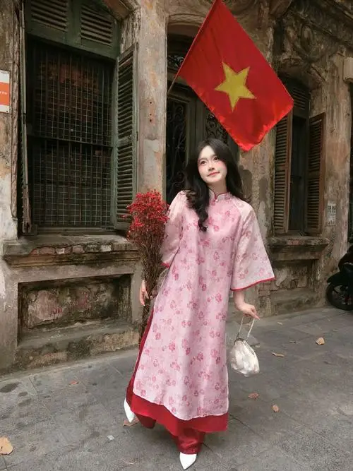 Bên hiên nhà treo cờ đỏ, người mẫu áo dài hoa đứng thẳng, tay cầm hoa và mỉm cười trong không khí hoài cổ